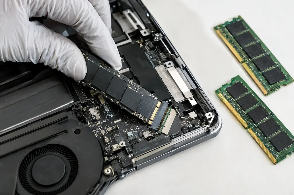 Gloved fingers seating an NVMe SSD into a laptop with new RAM sticks nearby